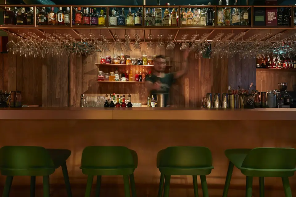 A bartender working behind a wooden club bar, green stools along it.